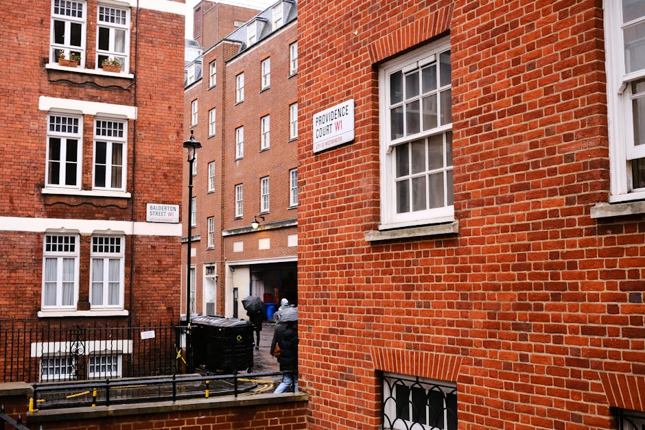 The image shows a narrow urban alleyway between red brick residential buildings, with a black wheelie bin placed against a short brick wall in the foreground. The bin has a yellow handle and appears t